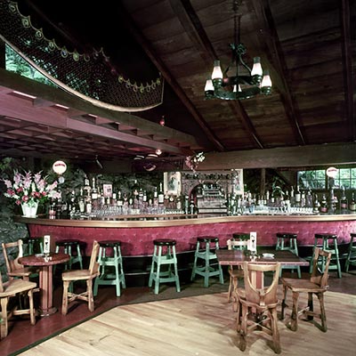 alt="A vintage photograph of the Rockroom Lounge at Shadowbrook Restaurant, featuring the natural rock hillside integrated into the interior wall, a stone fireplace, and mid-century modern lounge seating.