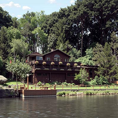 alt="A vintage evening view of Shadowbrook Restaurant from across Soquel Creek, with the multi-level building’s warm lights reflecting on the water and the red cable car visible on the hillside.