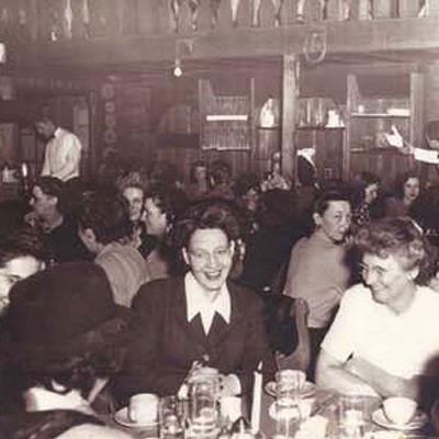 Historic archival photo of a social luncheon at Shadowbrook; women in classic 1950s attire are seated in a sunlit dining room surrounded by timber beams and lush garden views.