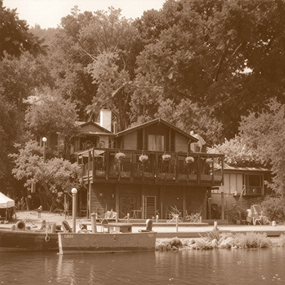 A vintage black-and-white photograph of the Shadowbrook Restaurant exterior in the mid-20th century, showing the original timber architecture and the beginning of its famous hillside gardens.