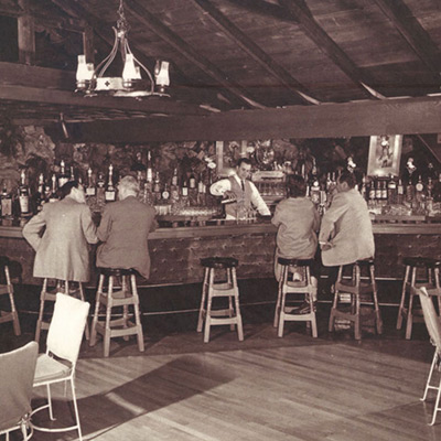 A vintage black-and-white photograph of the Rockroom Lounge at Shadowbrook, showing guests gathered around the natural rock walls and stone fireplace in a cozy, mid-century setting.