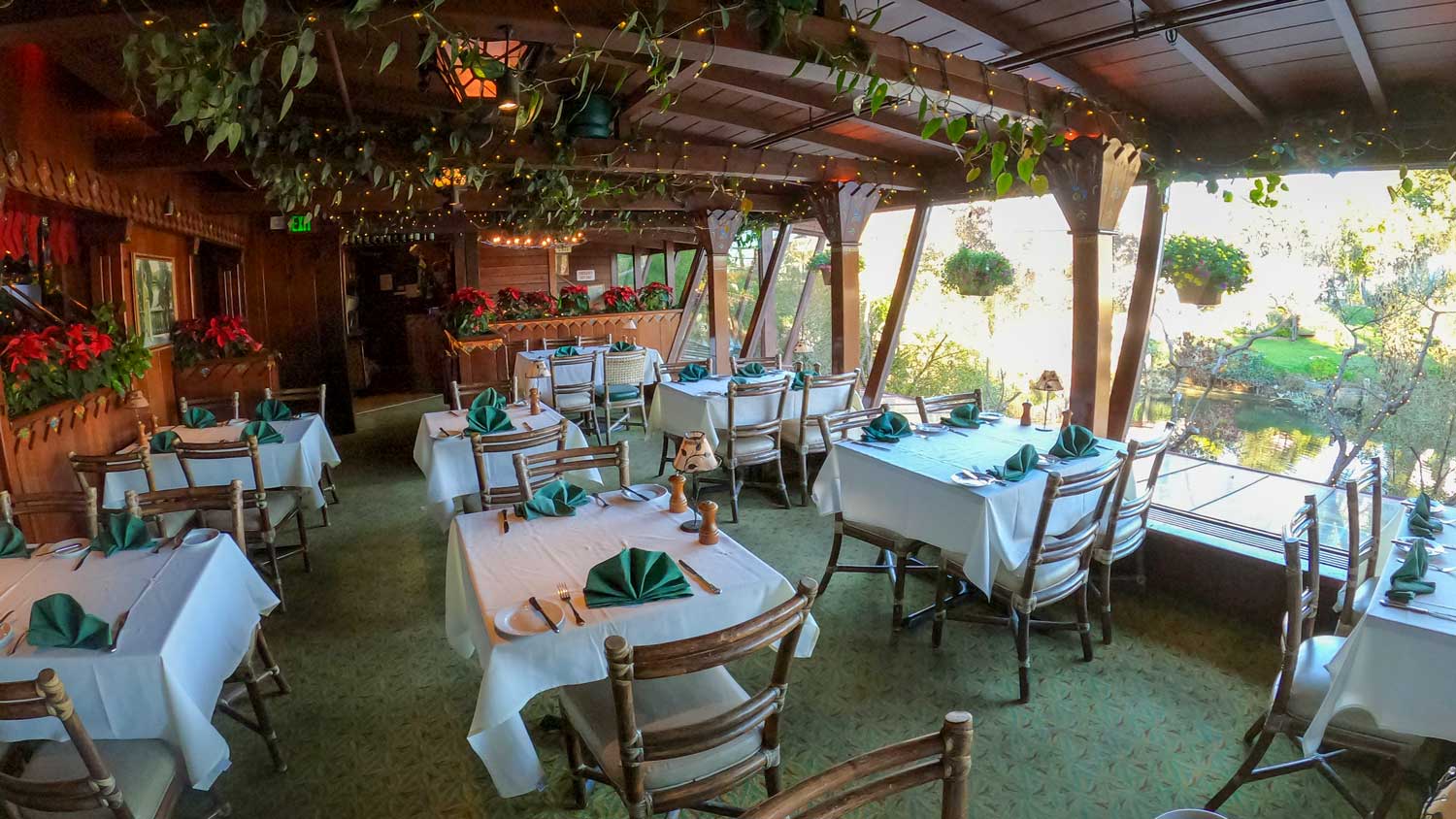 Interior dining room at Shadowbrook Restaurant, featuring candlelit wooden tables, white linens, and large windows overlooking the lush garden greenery.
